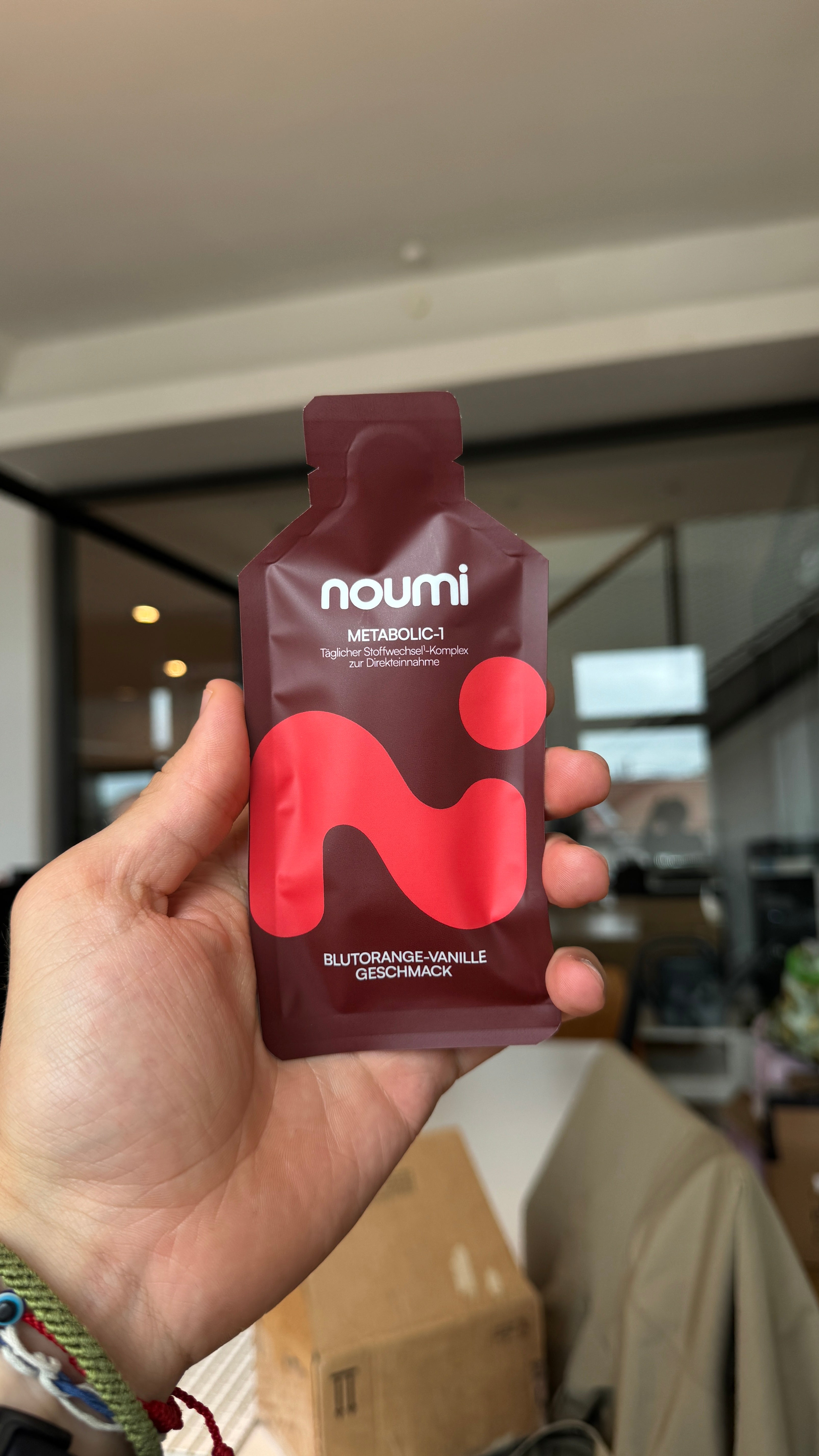 Hand hält ein noumi Metabolic-1 Sachet Blutorange-Vanille in einem Innenraum