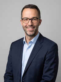 Lächelnder Mann mit Brille in dunklem Anzug und hellblauem Hemd vor grauem Hintergrund