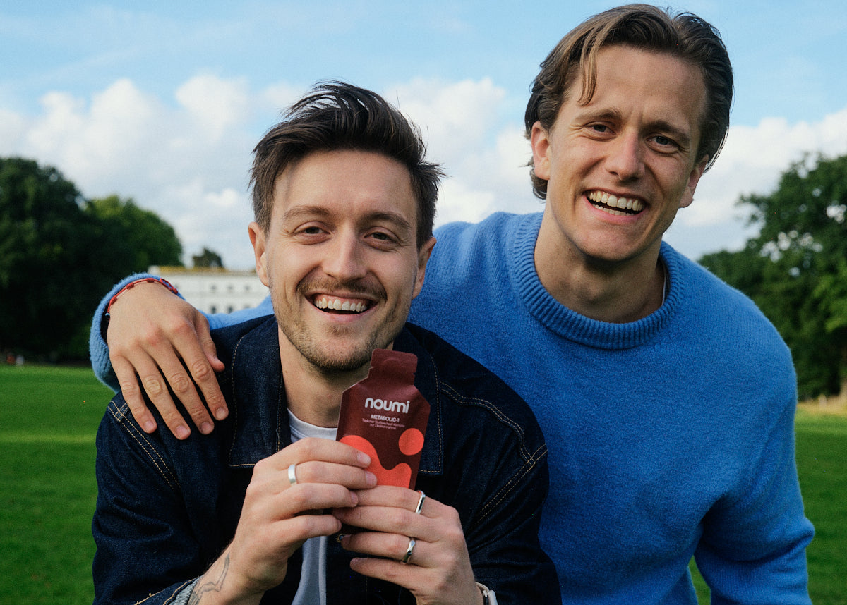 Zwei lächelnde Männer im Park, einer hält ein noumi Metabolic-1 Sachet in den Händen