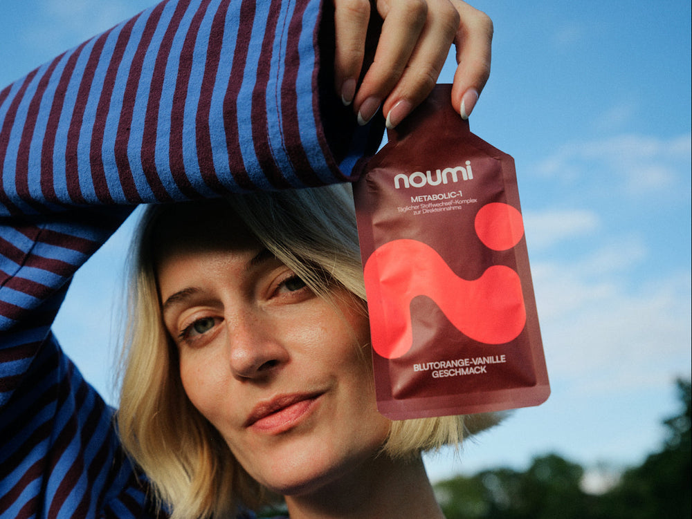 Frau hält ein noumi Metabolic-1 Gel-Sachet vor blauen Himmel in die Kamera
